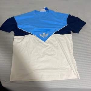 Säljer en snygg t-shirt från Adidas i blått och vitt. Den har en cool design med ett stort blått fält upptill och ett vitt fält nedtill, samt Adidas-loggan i vitt på bröstet. Ärmarna är korta och t-shirten är perfekt för en avslappnad stil.