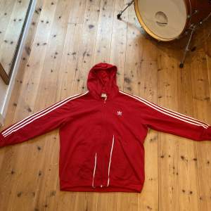 Säljer en snygg röd hoodie från Adidas med de klassiska tre vita ränderna längs ärmarna. Den har en dragkedja framtill och de går att juster både mudden och huvan  med snören (se bild 4/5. Perfekt för en avslappnad stil eller träning. Den är i bra skick och superbekväm!