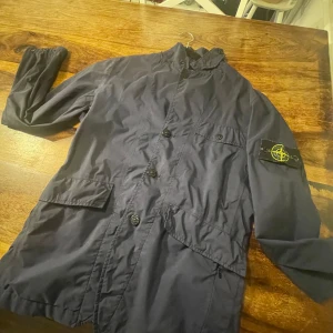 Mörkblå jacka från Stone Island - Säljer en snygg mörkblå jacka från Stone Island i mycket bra skick. Jackan har långa ärmar och knappar framtill. Perfekt för höst och vår med sin stilrena design och bekväma passform. Märkeslogga på ärmen ger en extra touch av stil.