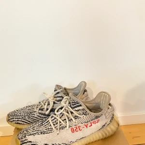 Adidas Yeezy Boost 350 V2 Zebra - Säljer ett par Adidas Yeezy Boost 350 V2 i Zebra-mönster. Skorna har en vit och svart randig design med röd text på sidan. De är i bra skick med lite slitage på sulorna. Perfekta för både vardag och fest!