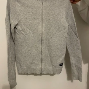 Grå kofta från Jack & Jones - Säljer en stilren grå kofta från Jack & Jones i storlek XS. Den har en praktisk dragkedja framtill och är tillverkad i ett mjukt material som passar perfekt för både höst och vår. Koftan har långa ärmar och en ribbad kant vid ärmslut och nederkant. Kontakta för fler bilder, lägre pris vid direktköp
