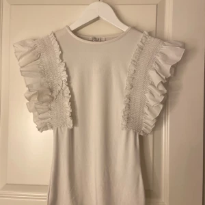 Vit topp med volangärmar - Säljer en superfin vit topp med volangdetaljer på ärmarna. Perfekt för sommardagar eller en festlig kväll. Toppen är i ett mjukt material och har en normal passform. Den är både stilren och söt, vilket gör den lätt att matcha med olika outfits.