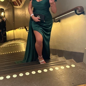 Grön långklänning med slits - Elegant grön långklänning i siden med en djup slits framtill. Klänningen har en offshoulder-design och en vacker drapering som ger en smickrande passform. Perfekt för festliga tillfällen! Har för mig att det förekommer en fläck vid släpet, kan därför behövas tvättas