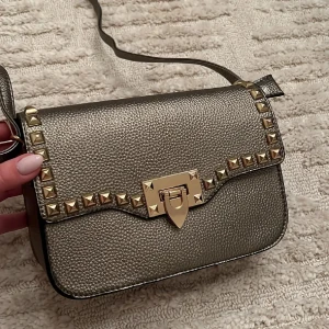 Snygg axelväska med nitar - Säljer en supercool axelväska i metalliskt grått med guldnitar och ett snyggt spänne framtill. Väskan har en justerbar axelrem och ett praktiskt innerfack med dragkedja. Perfekt för en utekväll eller när du vill lägga till lite edge till din outfit!