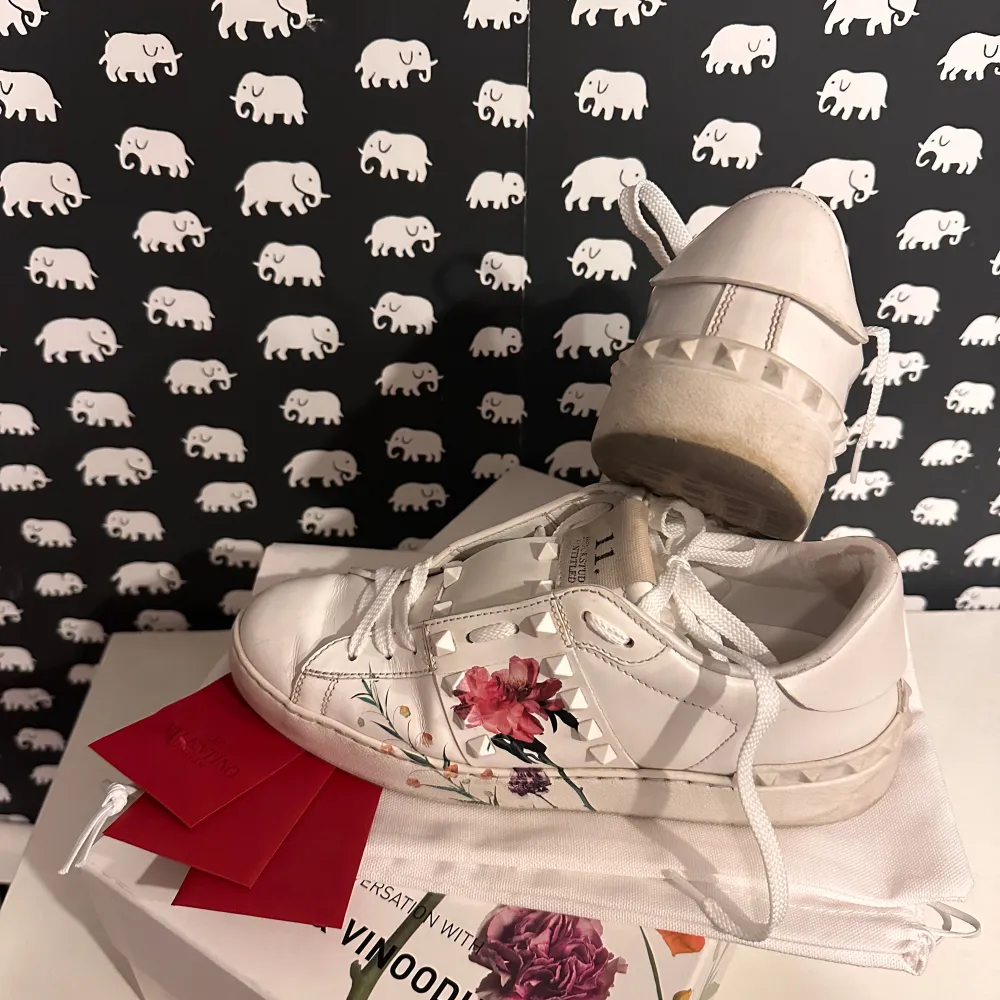 Säljer ett par snygga vita sneakers från Valentino i storlek 38. De har ett coolt blommönster på sidan och klassiska rockstud-detaljer. Perfekta för att ge din outfit en unik touch! Skorna är i mycket bra skick och kommer i originalförpackning. Collab med Inez & Vinoodh. Kvitto finns ej då det var en present. Nypris 7075kr.. Skor.