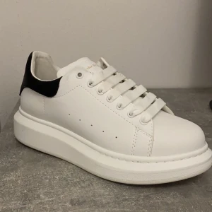  Alexander McQueen skor - Säljer ett par stilrena vita sneakers från Alexander McQueen med svart häl och guldfärgad logga. Skorna har en chunky sula och klassisk snörning. Perfekta för både vardag och fest! 🖤