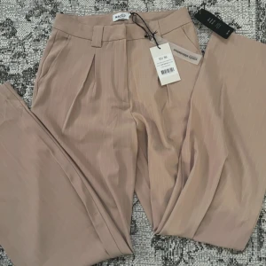 Beige kostymbyxor från NA-KD - Säljer ett par snygga beige kostymbyxor från NA-KD i storlek EU 36. Byxorna har en elegant randig design och är perfekta för både kontoret och festliga tillfällen. De är högmidjade med en klassisk passform och har knappar och dragkedja framtill. Helt nya med taggar kvar!