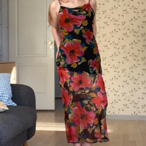 Blommig klänning från Urban Outfitters - Säljer en superfin blommig klänning från Urban Outfitters i storlek S. Den har ett färgglatt mönster med stora blommor i rosa, orange och grönt på en svart bakgrund. Klänningen är ärmlös med smala axelband och har en lång längd. 