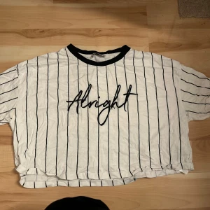 Vit och svart randig t-shirt från FB Sister - Säljer en vit kroppad t-shirt från FB Sister med svarta ränder och texten 'Alright' på framsidan. Den har en avslappnad passform och är kortärmad med en svart krage. Perfekt för en casual look!