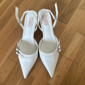 Vita pumps med klack och spänne - Säljer ett par eleganta vita pumps i syntetmaterial med spetsig tå och klack. De har snygga spännen och en stilren design, perfekt för fest eller bal. Skorna är i bra skick med några mindre märken på tån. 