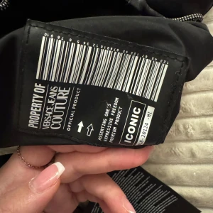 Svart ryggsäck från Versace Jeans Couture - Säljer en snygg svart ryggsäck från Versace Jeans Couture. Den har ett stort vitt logotryck på framsidan och praktiska dragkedjor. Perfekt för både vardag och speciella tillfällen. Ryggsäcken är i mycket bra skick och redo för nya äventyr!