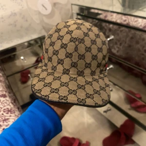 Beige keps med monogrammönster - Snygg beige keps från Gucci med det klassiska GG-monogrammet över hela ytan. Kepsen har en röd och grön randdetalj på sidan, vilket ger den en extra touch av stil. Perfekt för att lyfta vilken outfit som helst!