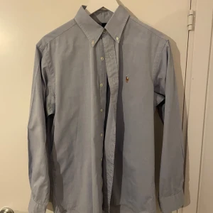 Ljusblå skjorta från Ralph Lauren - Säljer en klassisk ljusblå skjorta från Ralph Lauren i 100% bomull. Skjortan har en stilren design med knappar framtill och en liten broderad logga på bröstet. Perfekt för både vardag och fest! Passar bra till jeans eller chinos för en snygg look. 🕺
