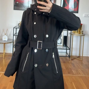 Svart trenchcoat från Calvin Klein - Säljer en snygg svart trenchcoat från Calvin Klein. Den har en stilren design med silverfärgade knappar och dragkedjor. Jackan har ett bälte i midjan med ett spänne som också har Calvin Klein-loggan. Perfekt för höst och vår!