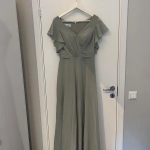 Balklänning  - En vacker klänning i en mjukt grågrön färg med en v-ringning och korta, rufsade ärmar. Klänningen har en smickrande passform och en elegant silhuett. Den är perfekt för en festlig tillställning eller en kväll ute.  Använd 1 gång!