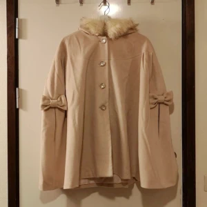 Beige poncho med fuskpäls & rosetter - Beige poncho/kappa med fluffig fuskpäls runt kragen. Det var svårt att få en consistent färg på den, så det varierar lite på bilderna ^^' Använd en gång. Har varit listad en gång innan hehe.. Köpare står för frakt om inget annat anges