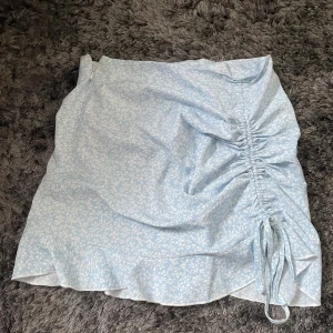 Ljusblå blommig kjol från Shein - Säljer en superfin ljusblå kjol från Shein med ett sött blommönster. Kjolen har en snygg rynkdetalj på sidan med knytband som ger en extra touch. Perfekt för sommardagar eller en avslappnad fest. Materialet känns lätt och bekvämt. Passa på att fynda denna sommarfavorit! 🌸