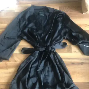 Säljer en elegant svart kimono i satin. Den har trekvartsärmar och ett knytband i midjan för en justerbar passform. Perfekt för en avslappnad hemmakväll eller som en stilfull detalj över en klänning.