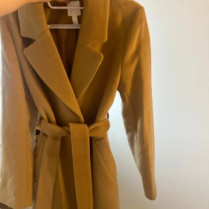 Beige/brun kappa med band  - En helt oanvänd kappa från H&M köpt förra säsongen. Kommer med band och kan tilläggas päls till. Har fickor och är ungefär 81 cm i längd från axlarna. Kan skickas fler bilder efter begäran! 