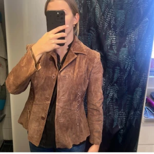 Brun mockajacka från BigL - Säljer en snygg brun mockajacka från BigL i storlek 38. Jackan har en klassisk design med knappar framtill och dekorativa snörningsdetaljer på sidorna. Perfekt för höst och vår, och ger en stilren look till vilken outfit som helst.