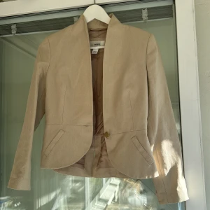 Beige kavaj från MNG - Säljer en snygg beige kavaj från MNG (Mango) i storlek XS. Den har en elegant design med en knapp framtill och långa ärmar. Perfekt för både vardag och festliga tillfällen. Kavajen är i bra skick och har en stilren look som passar till det mesta.