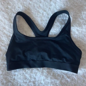 Svart sport-bh - Säljer en svart sport-bh i syntetmaterial. Den är perfekt för träning med sin tighta passform och bekväma design. Den har breda axelband och en elastisk kant nedtill för extra stöd.