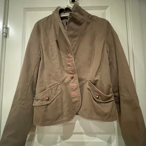 Beige kappa från H&M - Säljer en snygg beige kappa från H&M i storlek 38. Den har en stilren design med knappar framtill och på ärmsluten. Perfekt för höst och vår, och funkar både till vardags och fest.