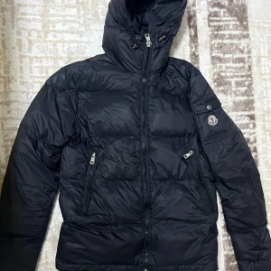 Svart pufferjacka från Moncler - Säljer en snygg svart pufferjacka från Moncler. Jackan har en dragkedja framtill och praktiska fickor med dragkedjor. Den är perfekt för kalla dagar med sin varma och vadderade design. Inuti finns en cool serietidningsdetalj som ger jackan en unik touch. Perfekt för vintern!