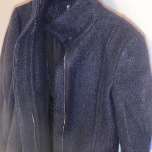 Mörkblå ullkappa med dragkedja - Säljer en stilren mörkblå kappa i ull (teddytyg)  med dragkedja framtill. Den har en hög krage och långa ärmar, perfekt för kyliga höst- och vinterdagar. Kappan är helt oanvänd, nypris 1500kr