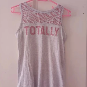Grå ärmlös topp med text - Säljer en grå ärmlös topp med texten 'TOTALLY' i glittriga rosa bokstäver. Toppen har ett coolt mönster i mesh upptill som ger en extra touch. Perfekt för sommardagar eller en avslappnad fest!