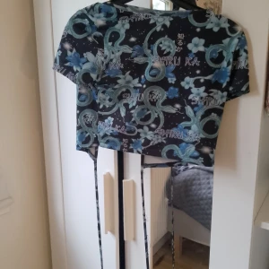 Mönstrad kortärmad topp med drakar - Säljer en cool kortärmad topp med ett unikt mönster av drakar och blommor i blått och grönt på en svart bakgrund. Toppen har en öppen rygg med knytband som ger en snygg detalj. Perfekt för en festlig kväll eller en dag när du vill sticka ut! har dok två små hål men dom syns inte 