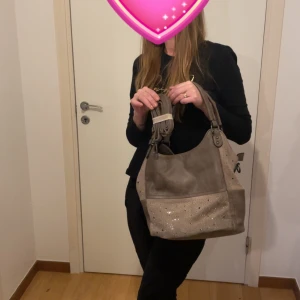 Mandoline Beige axelväska med glittriga detaljer - Snygg beige axelväska med glittriga detaljer och justerbara axelremmar. Väskan har en praktisk dragkedja och är perfekt för att bära med sig det viktigaste. Den stilrena designen gör den lätt att matcha med olika outfits.