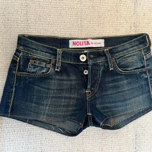 Mörkblå Lågmidjade jeansshorts från Nolita - Snygga mörkblå jeansshorts från Nolita med coola nitar på bakfickorna. Shortsen har en lågmidjad passform och klassisk femficksdesign med dubbla knappar fram. Perfekta för en avslappnad stil! Säljer bara för att dom är för små!