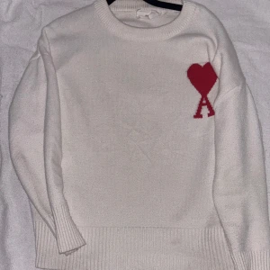 Vit stickad tröja från Ami - Säljer en stilren vit stickad tröja från Ami med ett rött hjärta och bokstaven 'A' på bröstet. Tröjan har långa ärmar och ribbade kanter vid ärmslut och nederkant. Perfekt för en trendig och bekväm look.