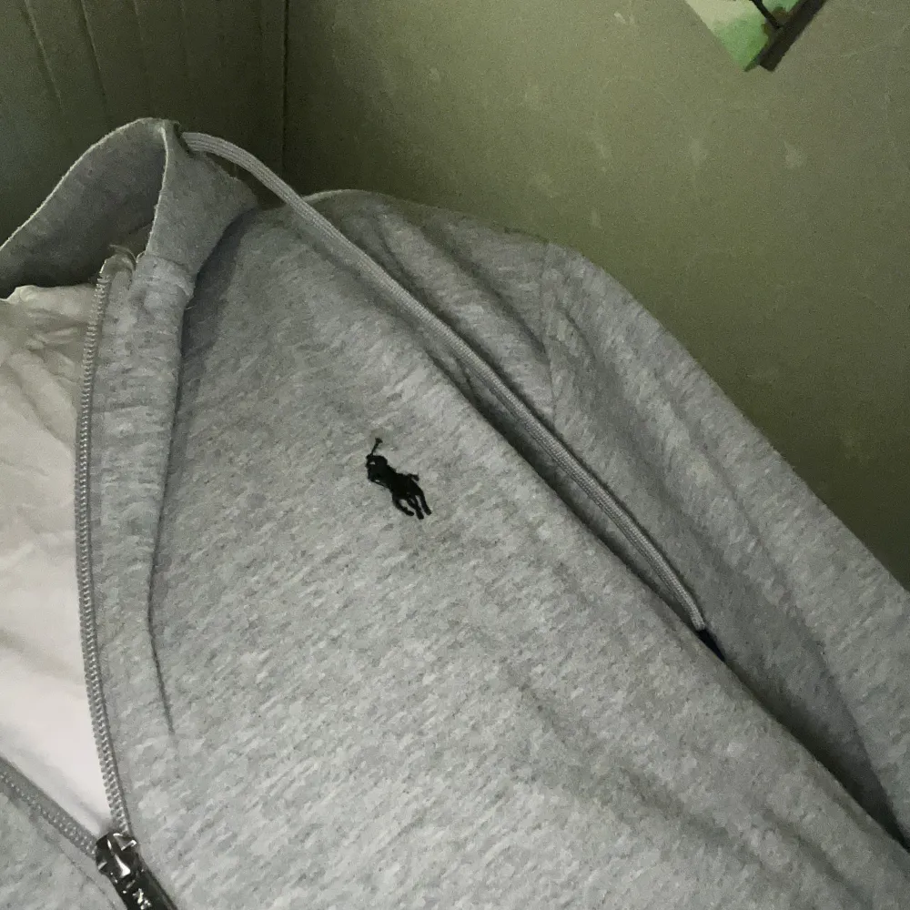 Säljer en stilren grå hoodie från Ralph Lauren med dragkedja och justerbar huva. Perfekt för en avslappnad look. Den har en liten logotyp broderad på bröstet. Passar bra till både jeans och joggers.. Neuletakit & Villapaidat.