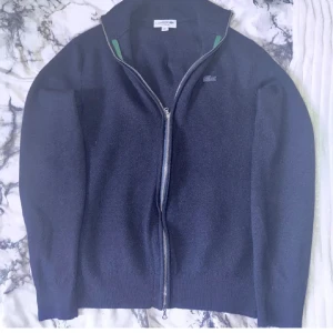 Mörkblå ulltröja från Lacoste - Säljer en stilren mörkblå tröja från Lacoste i 100% ull. Tröjan har en dragkedja framtill och en diskret logga på bröstet. Perfekt för kyligare dagar med sin mjuka och varma känsla. Passar bra till både jeans och chinos.