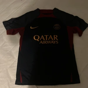 PSG träningströja - Snygg svart och röd fotbollströja från Nike med korta ärmar. Tröjan har en gul Nike-logga och Qatar Airways som sponsortryck på framsidan. Perfekt för fotbollsfans som vill visa sitt stöd.Nästan aldrig använd