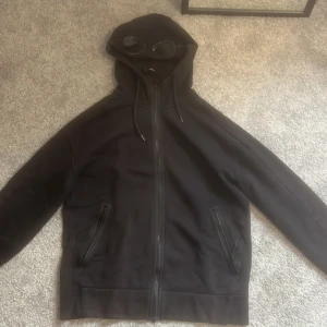 C.P. Company - Säljer en svart hoodie från C.P. Company med inbyggda glasögon i huvan. Den har dragkedja framtill och två fickor med dragkedjor. Perfekt för en unik och stilren look. Väl använd och slitningar finns (fråga om bild ifall du vill se bättre). Solblekningen börjar ta sig.