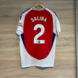 Röd och vit fotbollströja från Adidas - Snygg röd och vit fotbollströja från Adidas med 'Emirates Fly Better' tryckt på framsidan. Tröjan har korta ärmar och ett stilrent mönster med marinblå detaljer. Namnet 'Saliba' och nummer 2 pryder baksidan. Perfekt för fotbollsfans!