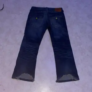 Säljer ett par blå jeans med slitningar på framsidan och slitna kanter vid bensluten. Jeansen har en klassisk femficksdesign och är tillverkade i denim. Perfekta för en avslappnad look. OBS: ACCEPTERA INTE TILLBAKA SKICKNINGAR! 