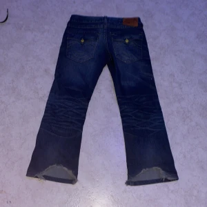 Blå jeans med slitningar - Säljer ett par blå jeans med slitningar på framsidan och slitna kanter vid bensluten. Jeansen har en klassisk femficksdesign och är tillverkade i denim. Perfekta för en avslappnad look. OBS: ACCEPTERA INTE TILLBAKA SKICKNINGAR! 