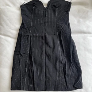 Svart axelbandslös klänning från Zara - Snygg svart axelbandslös klänning från Zara med diskreta vita ränder. Klänningen har en dragkedja i sidan för enkel påklädning och en elegant passform som framhäver figuren. Perfekt för en stilren look.