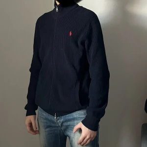 Mörkblå stickad tröja från Polo Ralph Lauren - Säljer en stilren mörkblå stickad tröja från Polo Ralph Lauren med dragkedja och det klassiska röda logotypbroderiet på bröstet. Jättebra skick 