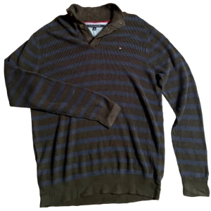 Randig tröja från Tommy Hilfiger - Snygg randig tröja från Tommy Hilfiger i mörkgrön och blå. Tröjan har långa ärmar och en unik krage med knappar. Perfekt för en stilren och avslappnad look.