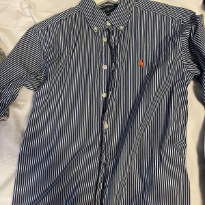 Blå och vit randig skjorta från Ralph Lauren - Snygg blå och vit randig skjorta från Ralph Lauren med klassisk krage och knappar framtill. Skjortan har en broderad orange logotyp på bröstet och är tillverkad i 100% bomull. Perfekt för en stilren look!