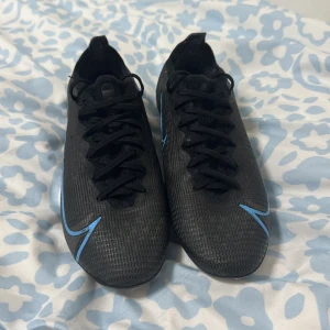 Nike mercurial elite vapor 16  - Snygga svarta Nike fotbollsskor med blå detaljer och snörning. Skorna har en slitstark sula med dobbar för bra grepp på planen. Perfekta för fotbollsspelare som vill ha stil och funktion.