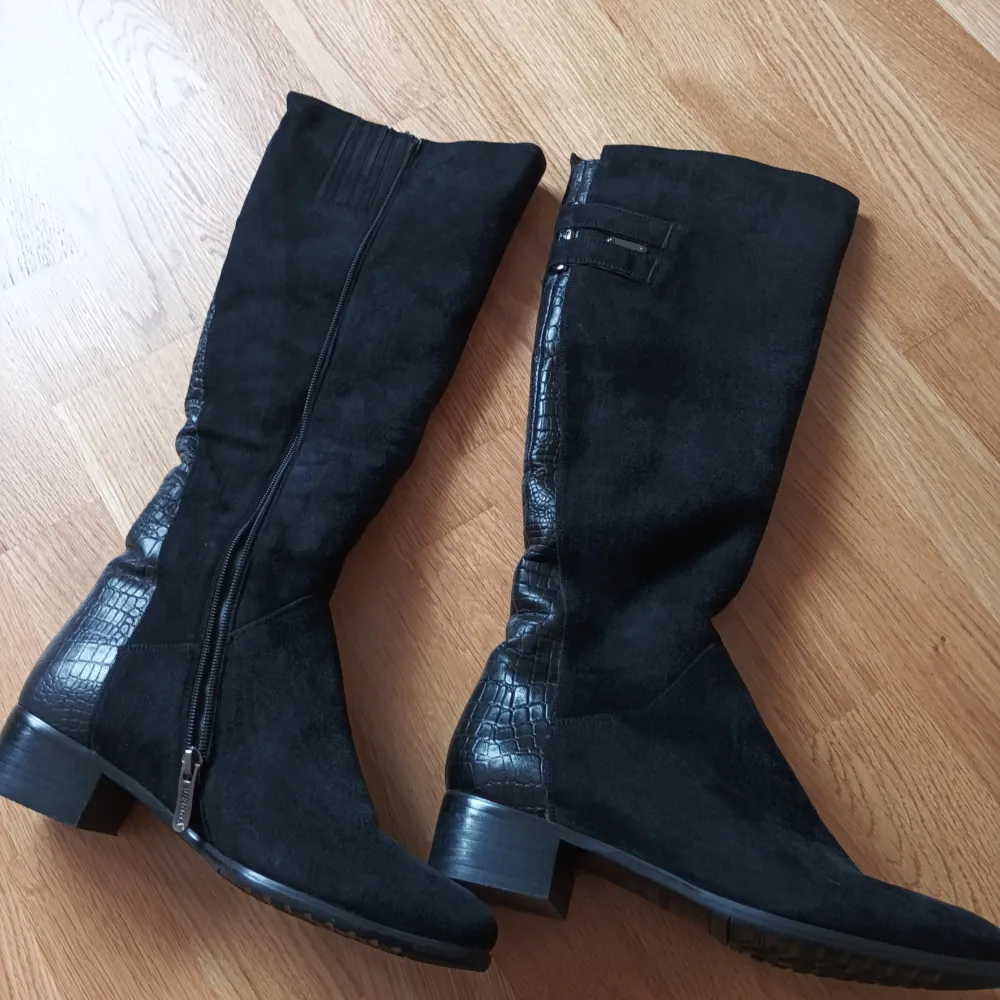 Snygga svarta stövlar i mocka med krokodilmönstrade detaljer och dragkedja på sidan.vinterstövlar i äkta läder med äkta päls inuti. Perfekta för en stilren look. De har en låg klack och är knähöga, vilket ger en elegant touch. . Kengät.