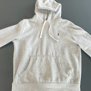 Grå hoodie från Polo Ralph Lauren - Säljer en stilren grå hoodie från Polo Ralph Lauren. Den har en klassisk design med en liten broderad logga på bröstet och en praktisk magficka. Huvan har en justerbar dragsko för extra komfort. Perfekt för en avslappnad look.