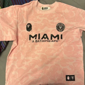 Rosa fotbollströja från A Bathing Ape - Säljer en rosa fotbollströja från A Bathing Ape i samarbete med Miami. Tröjan har ett unikt mönster och tryck med Miami och A Bathing Ape-loggor. Perfekt för dig som gillar streetwear och fotboll.