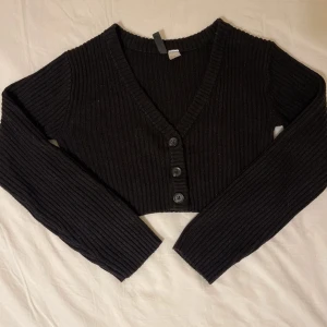 Svart ribbad kofta - Jag säljer en svart ribbad kofta med v-ringning och knappar framtill. Koftan är croppad och har långa ärmar.